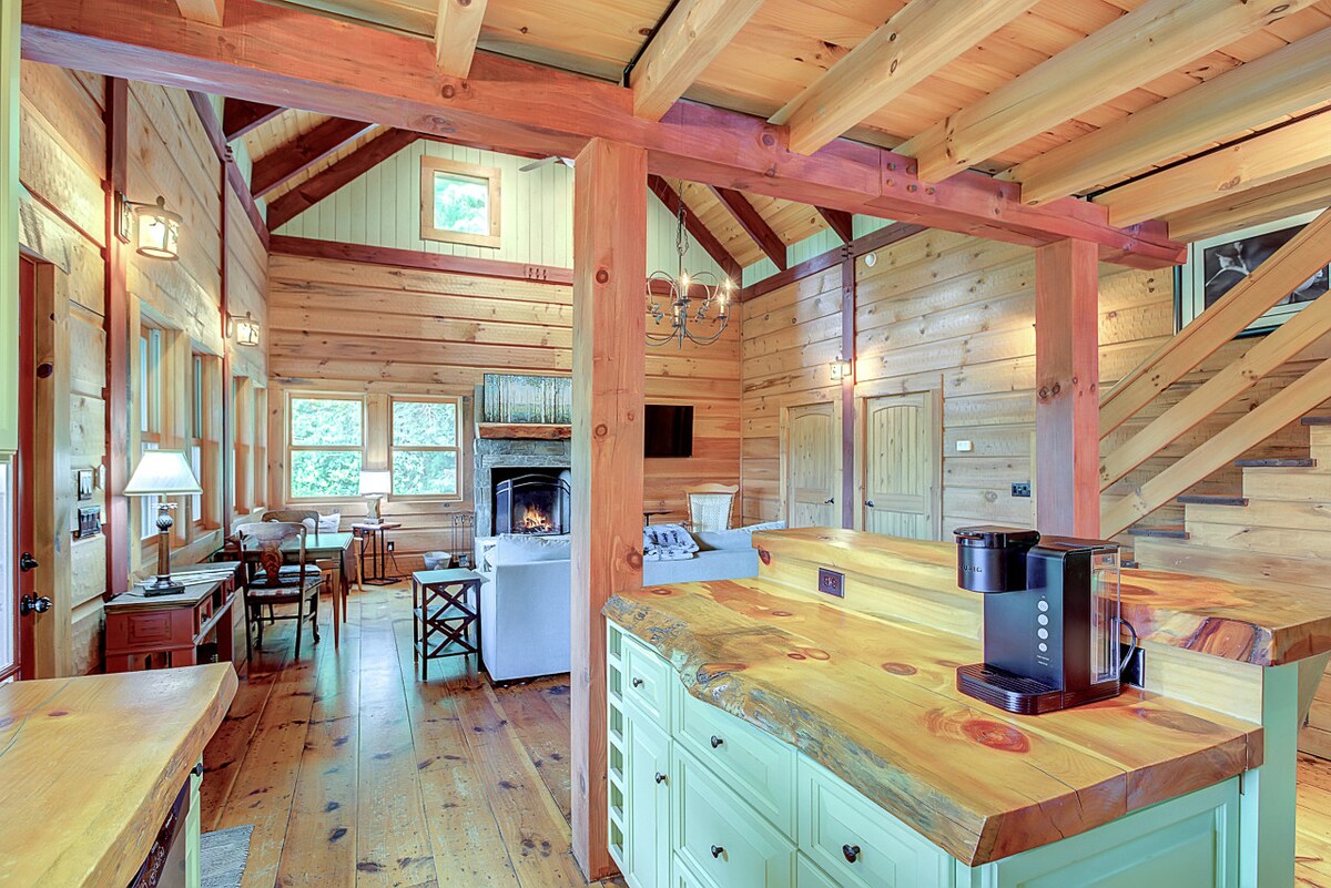 An open concept kitchen and living space features natural wood accents throughout the walls and ceiling. A spacious island with a unique wooden countertop complements the area. Large windows invite natural light, enhancing the cozy atmosphere next to the fireplace and dining space.