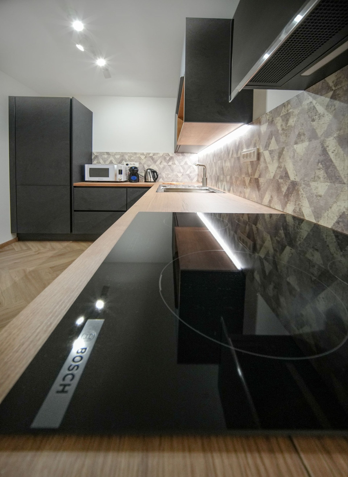 A modern kitchen features sleek black cabinetry and a stylish countertop. Integrated appliances, including a microwave and coffee maker, are neatly arranged, complemented by a geometric backsplash. Under-cabinet lighting enhances the workspace, reflecting off the smooth surface for a bright and functional cooking area.