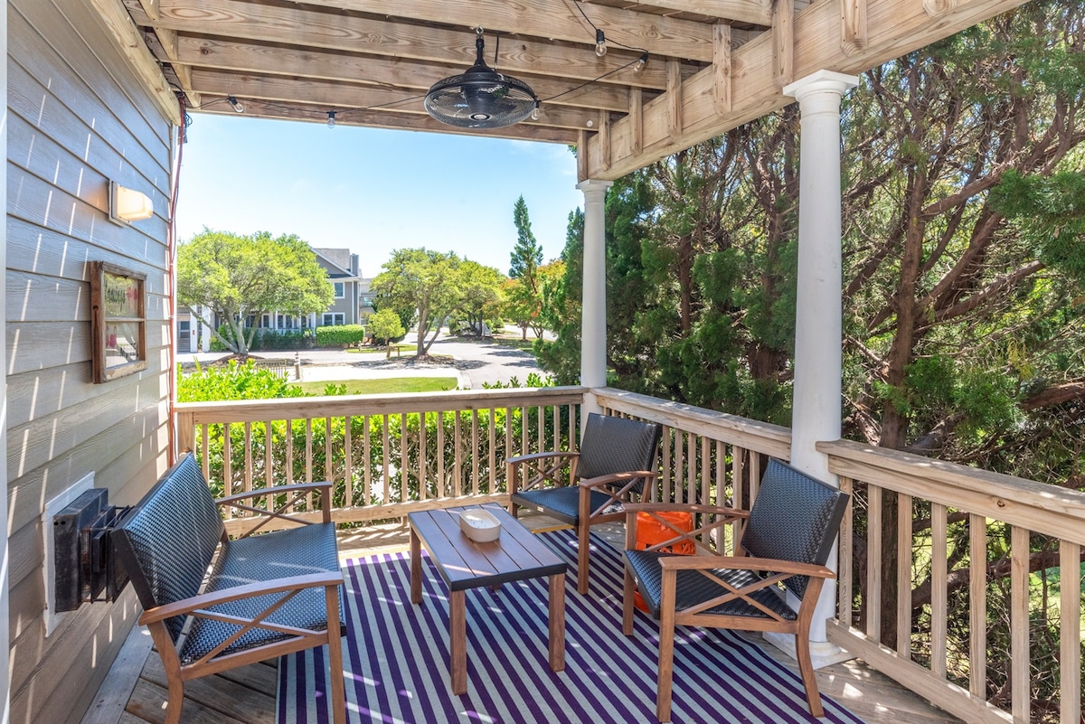 A welcoming outdoor deck area features four comfortable chairs arranged around a central table. A striped rug adds a touch of color to the space, complemented by the surrounding greenery and nearby buildings. A ceiling fan is visible above for comfort.