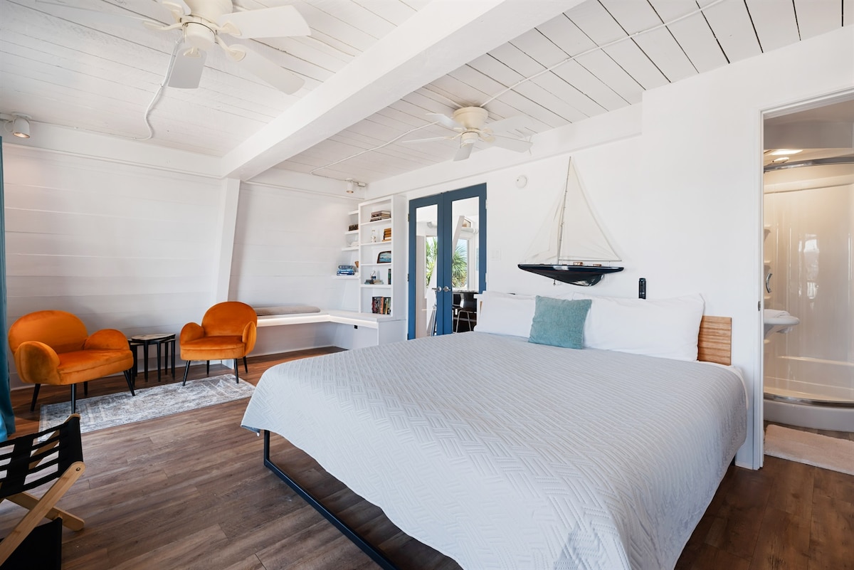 A spacious bedroom is presented, featuring a large bed with a textured white coverlet. Two comfortable orange chairs are positioned nearby, and a bookshelf with decorative items is visible. Bright light fills the room, with a small bathroom area seen in the background.
