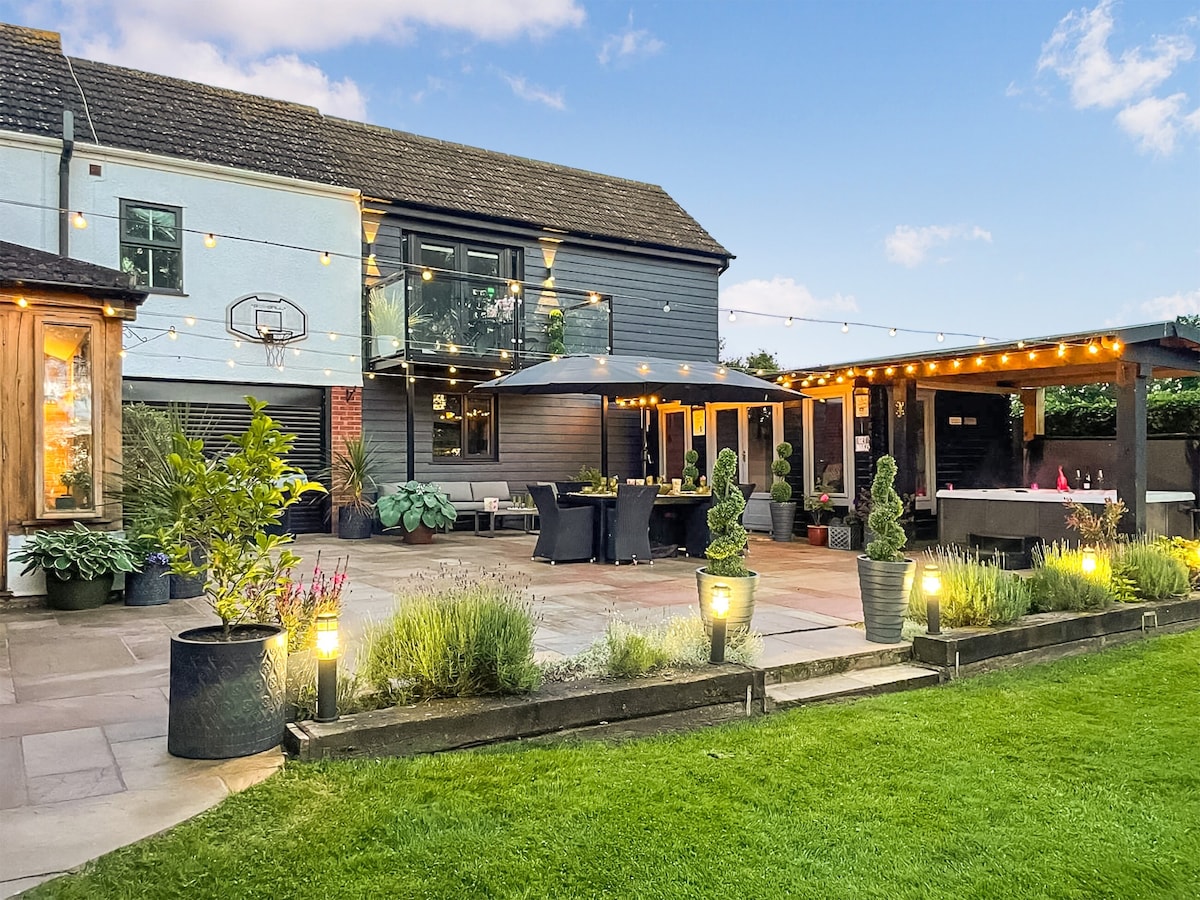 The outdoor area of the cottage features a spacious stone patio adorned with string lights and stylish landscaping. A large umbrella provides shade over a dining table, while the hot tub is positioned nearby, surrounded by greenery and decorative planters, creating an inviting atmosphere for relaxation.