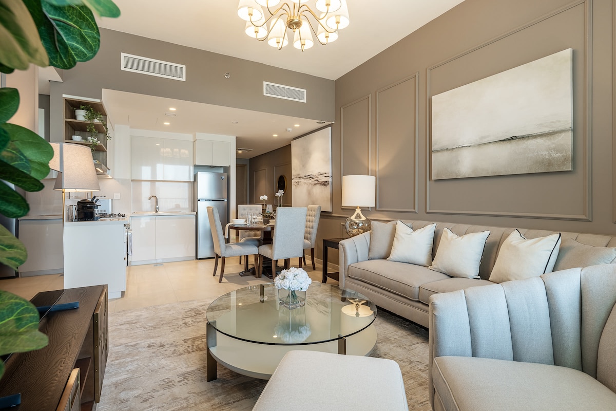 The living area features comfortable taupe couches surrounding a glass coffee table, with a plush rug beneath. A modern dining space with a round table and upholstered chairs is visible in the background, complemented by a well-equipped kitchen, highlighting an elegant atmosphere.