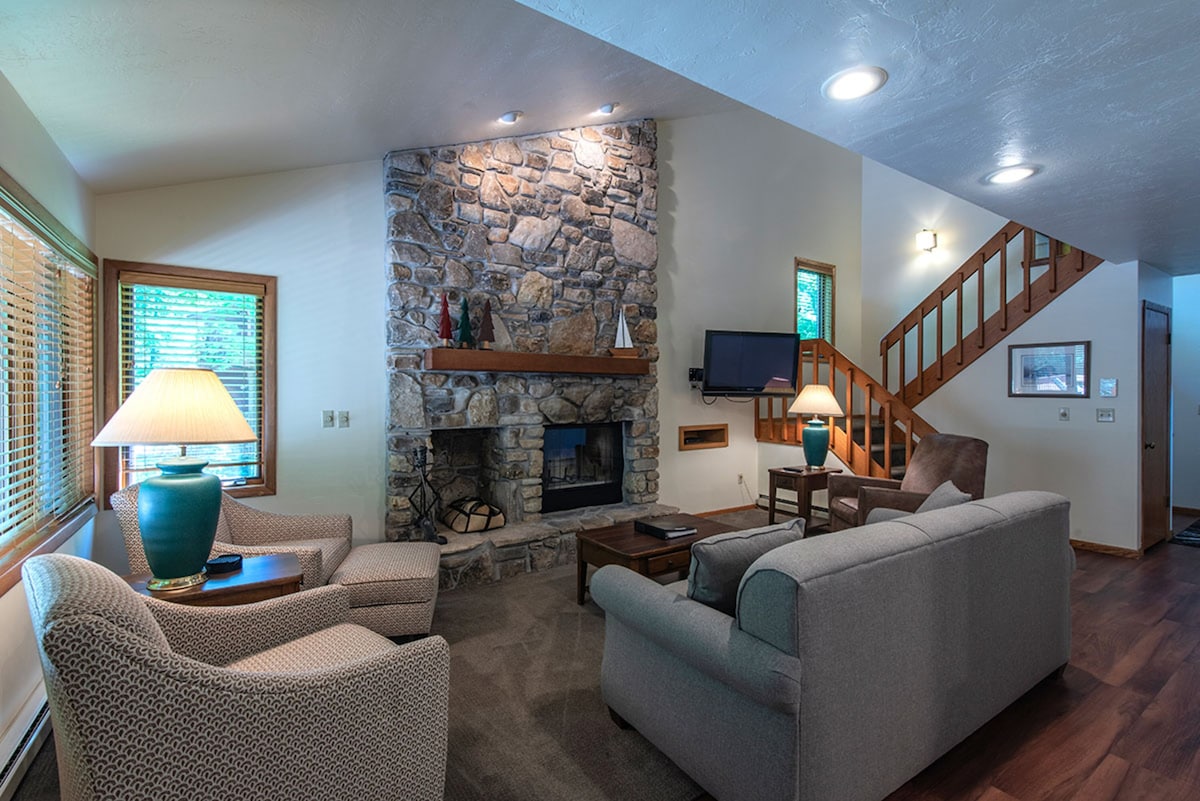 A spacious living area is highlighted by a stone fireplace, offering a cozy focal point. Two comfortable armchairs and a sofa provide ample seating, while a wooden staircase leads to the upper level. Large windows allow natural light to fill the room.