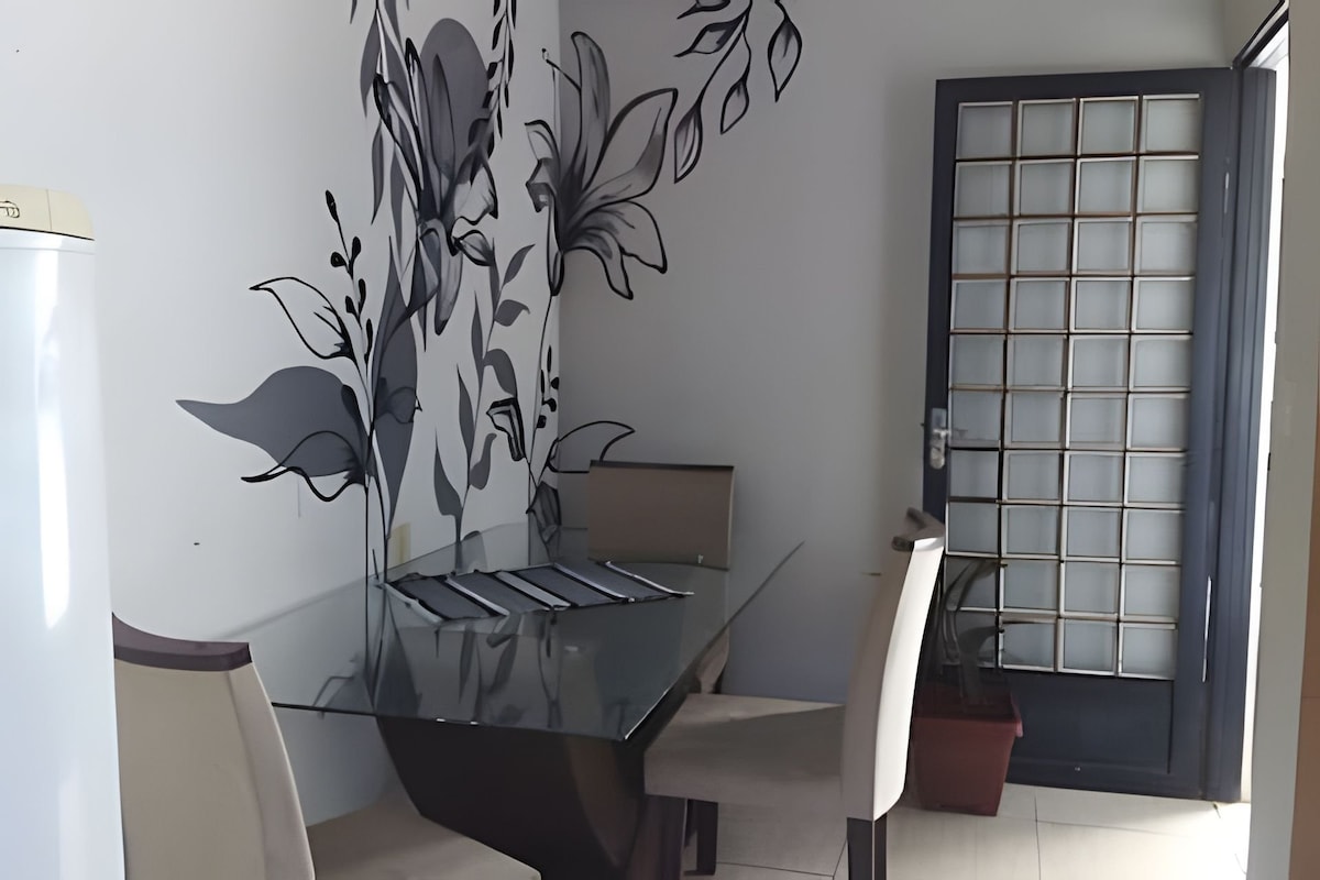 A dining area is featured, highlighted by a glass table surrounded by four light-colored chairs. A wall mural displaying floral designs adds visual interest. A door with a grid pattern provides access to the outside.