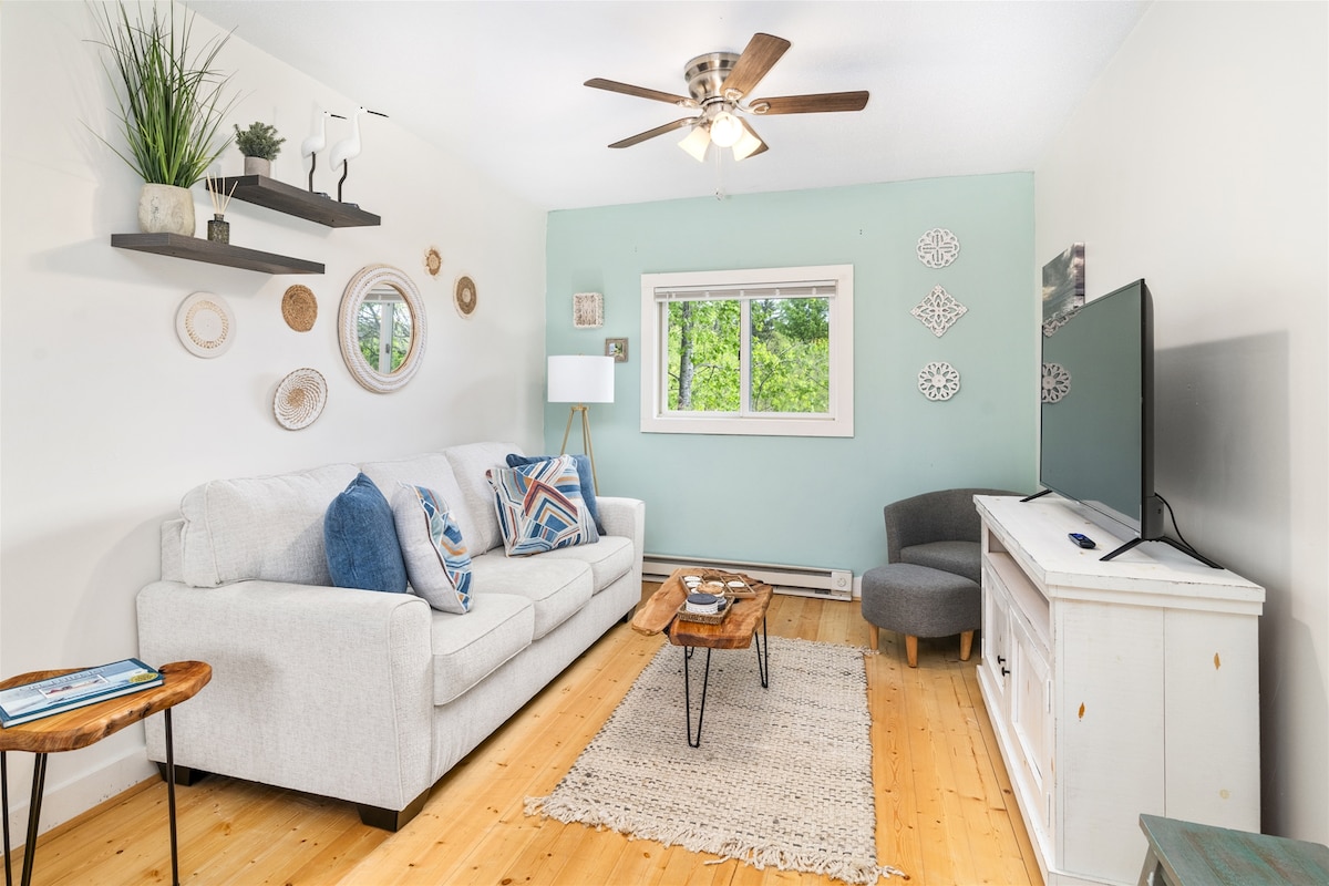 A light-filled living area features a comfortable sofa adorned with decorative pillows. A wooden coffee table sits beside the couch, with a textured area rug on the floor. A flat-screen TV is mounted on a white console, and woven wall decor adds visual interest.