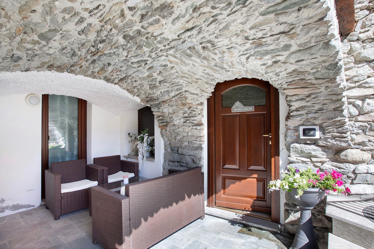 An entrance area features a stone archway, providing a rustic charm. Outdoor seating is arranged with a coffee table and wicker sofas, complemented by potted flowers, enhancing the welcoming appearance. The wooden door invites guests into the holiday apartment, suggesting ease of access.
