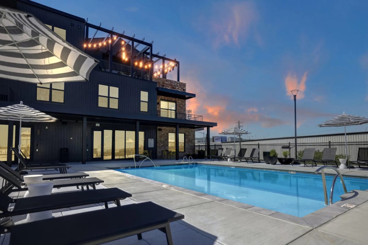 The communal pool area is surrounded by lounge chairs and shaded umbrellas, offering a space for relaxation. The sparkling water reflects the evening sky, with warm hues appearing as the sun sets over the elegant building in the background.
