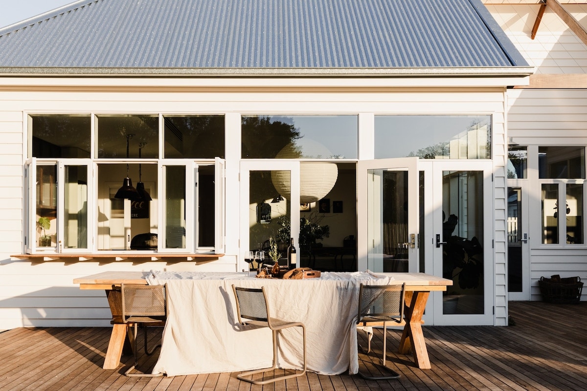 An expansive outdoor dining area features a large wooden table set with a neutral tablecloth. Surrounding the table are several metal chairs, with large windows allowing natural light to flood the space. The serene atmosphere is complemented by a lantern hanging overhead.