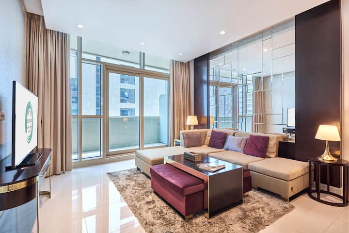 Dhomë e rehatshme ndenjjeje me një divan të madh modular, ndriçim të butë dhe dritare nga dyshemeja deri në tavan që hapen në një ballkon privat. Planimetria e ndritshme krijon një vend çlodhës për t’u çlodhur, për të parë televizor ose për të shijuar momente qetësie me dritën natyrale që mbush 