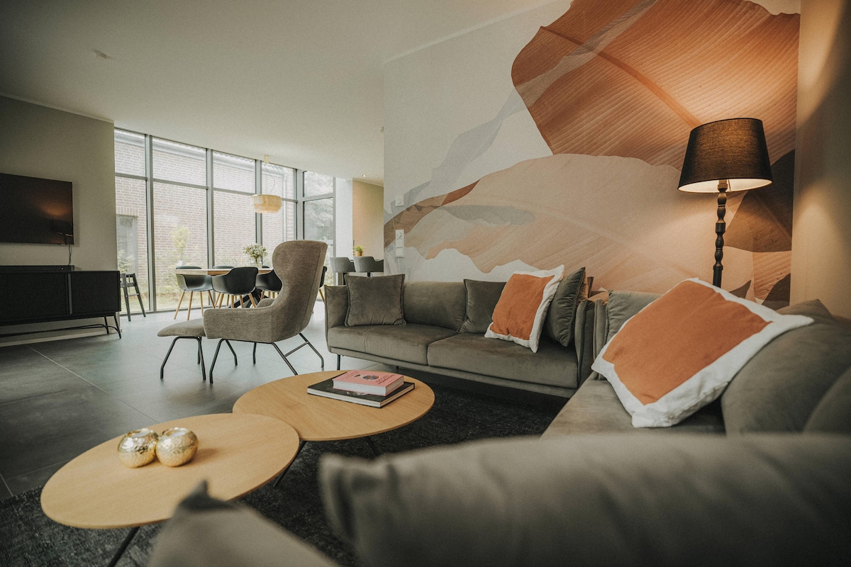 A modern living room features a comfortable sofa adorned with decorative pillows. A round coffee table holds stacked books and decorative objects. Large windows provide ample natural light, and a wall with artistic design adds a touch of sophistication to the space.