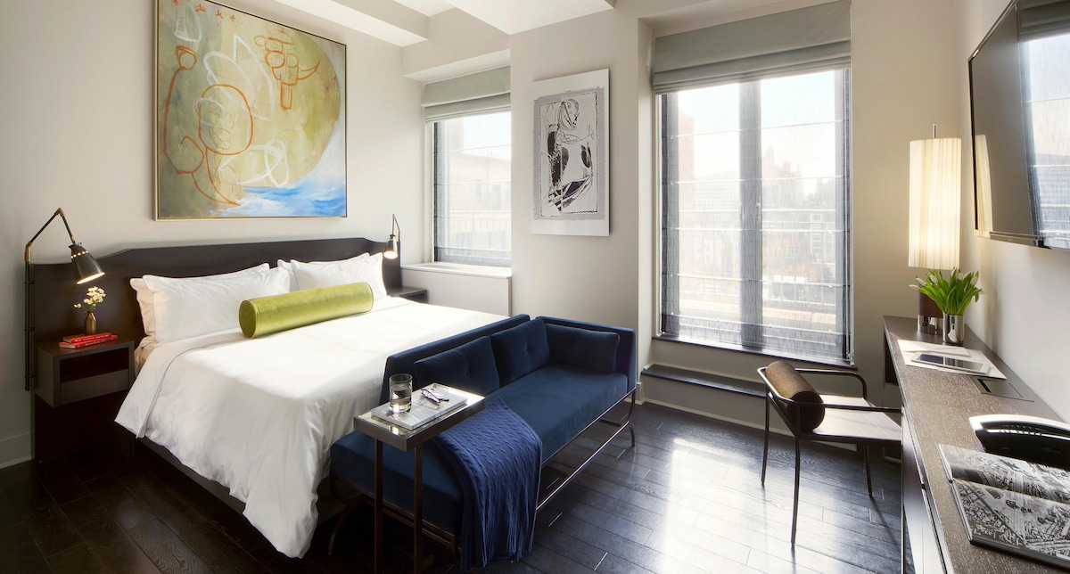 A spacious room features a large bed with crisp white linens and a decorative green bolster. A blue sofa and a small table are positioned nearby. Natural light streams through large windows, highlighting modern artwork on the walls and a sleek desk with a chair.