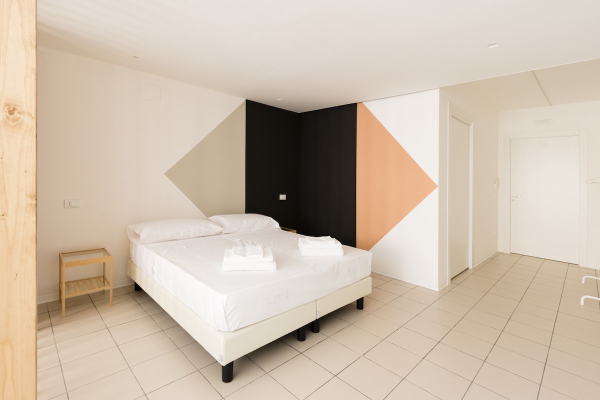 A spacious sleeping area showcases a double bed with neatly arranged linens. The minimalist decor features geometric wall designs in muted colors. A bedside table is positioned beside the bed, and natural light fills the room through a doorway visible in the background.