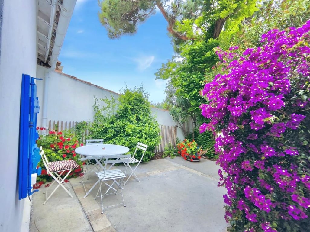 A serene outdoor patio area is surrounded by vibrant flowers and greenery. A round table with four chairs is positioned on a concrete surface, while colorful blossoms add charm to the space. The clear blue sky overhead complements the inviting atmosphere.