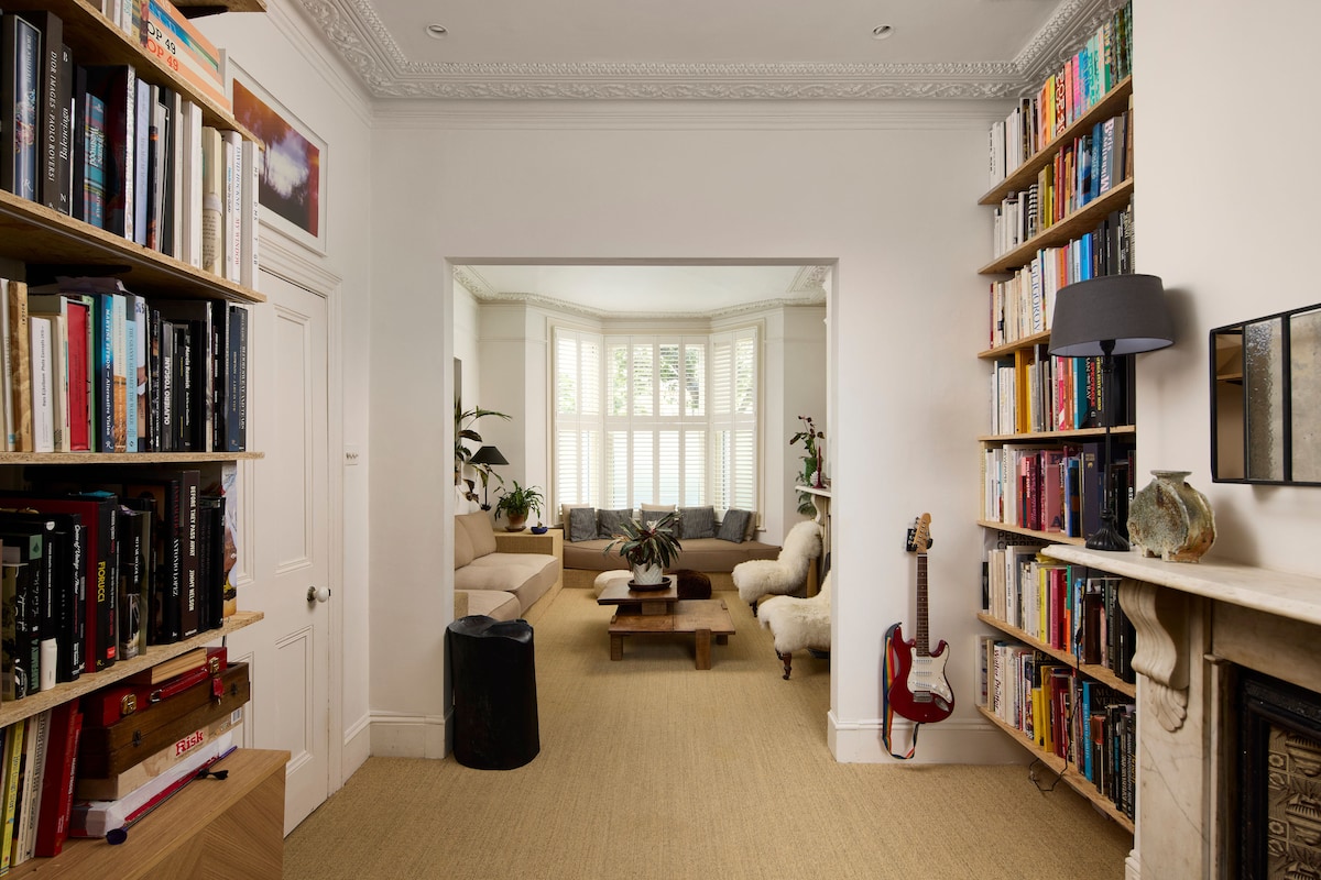 An inviting living room is highlighted by a cozy seating area and large windows allowing natural light to fill the space. Bookshelves line the walls, filled with a variety of books. A guitar leans against a table, adding a personal touch to the elegant decor.