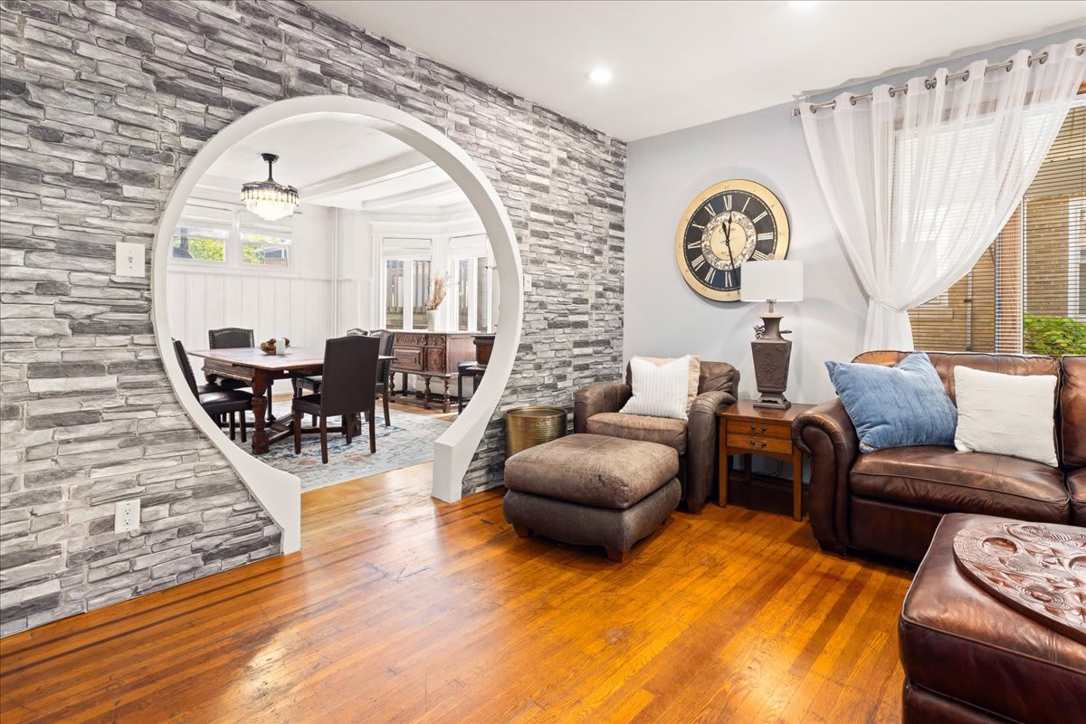 A cozy living area features a distinct archway leading to a dining space. Stone wall accents enhance the room, while warm wooden floors complement the comfortable seating arrangement, which includes a leather sofa and a plush chaise lounge. Natural light filters through sheer curtains.