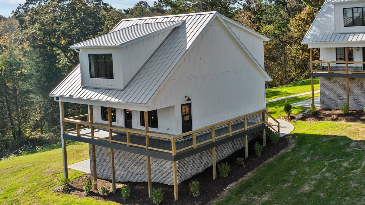 The exterior of a modern two-story cottage is showcased, featuring a white façade and a metal roof. The building is elevated on a deck supported by stone pillars, surrounded by lush greenery. Large windows allow for ample natural light, enhancing the inviting appearance.