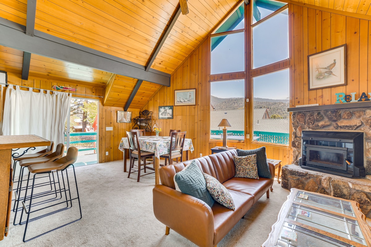 The living area features a warm and inviting design with a large, brown leather sofa and a wooden coffee table. A dining table with four chairs is present, along with a bar area featuring high stools. Large windows offer views of the outdoors, illuminating the space.