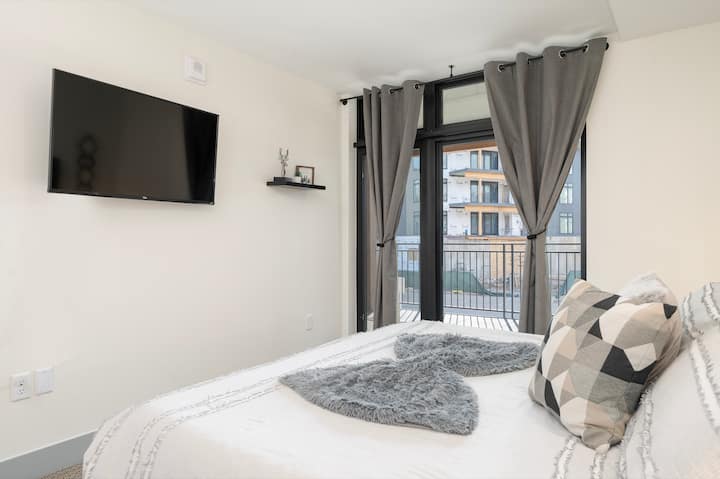 Een moderne slaapkamer met een aan de muur gemonteerde tv, een kingsize bed  en  grote ramen met uitzicht op een balkon.