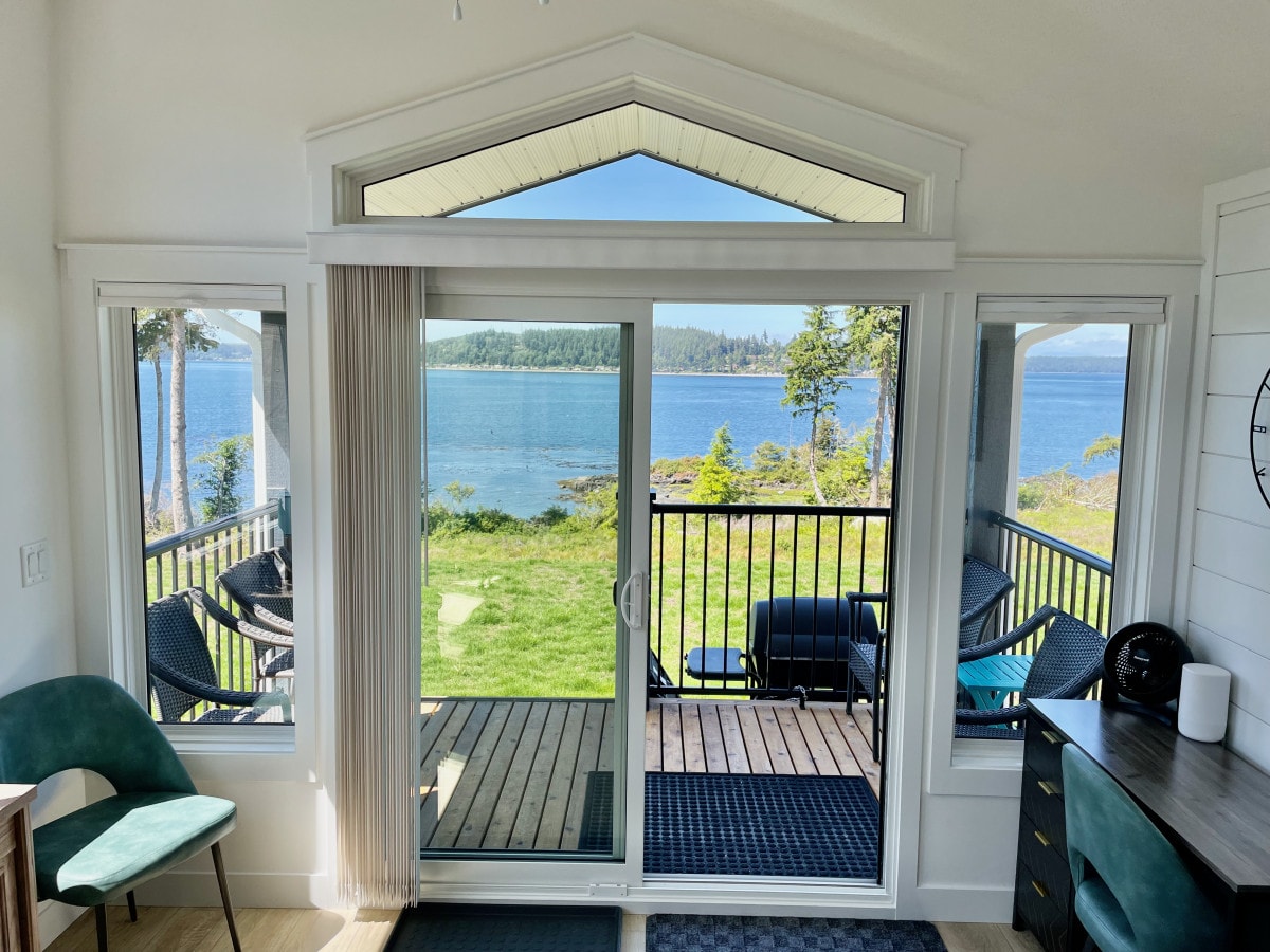 A large glass sliding door opens to a private deck, showcasing views of Johnstone Strait. The bright interior features large windows allowing natural light to fill the space, complemented by a small desk and cozy chair arranged near the entrance.