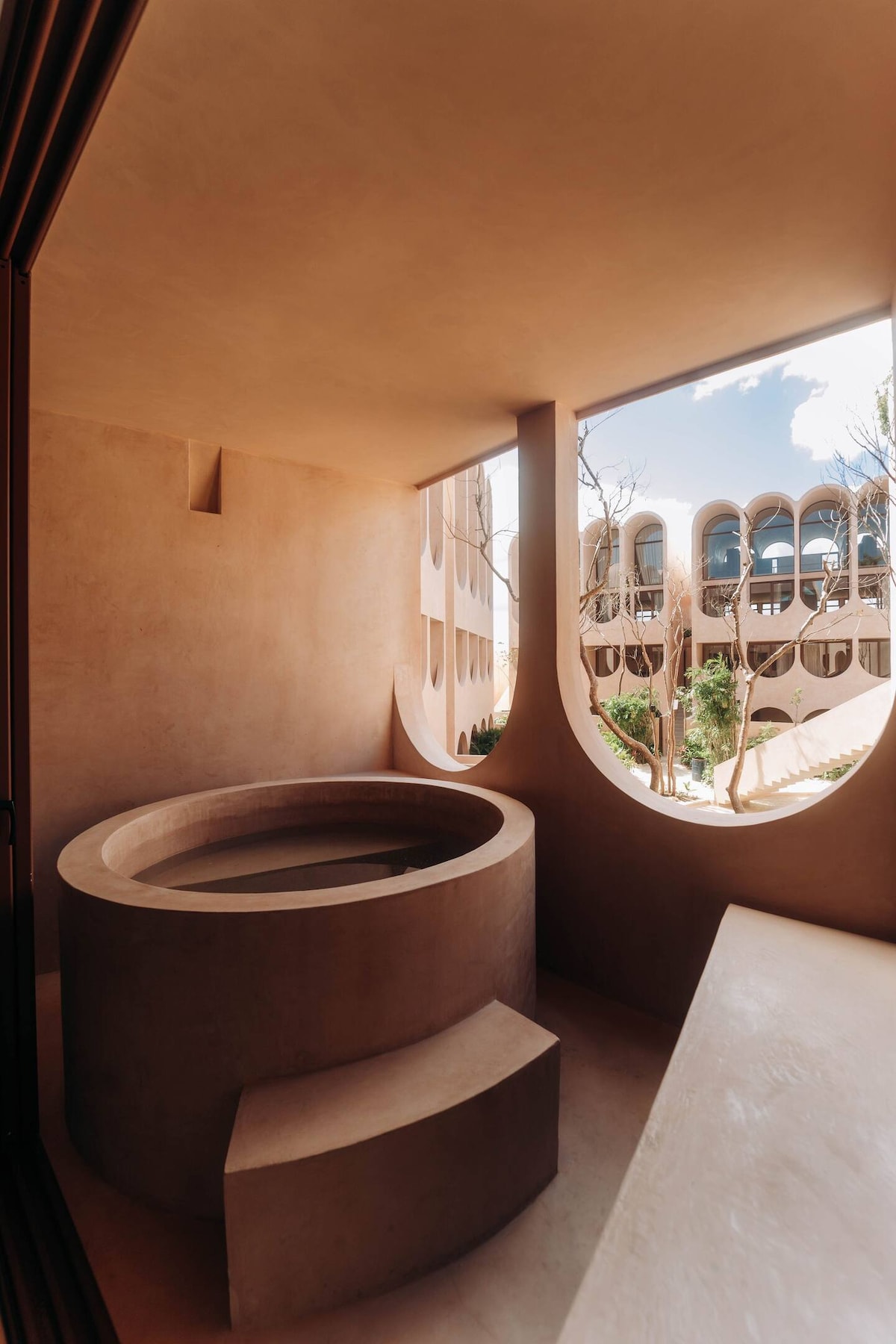 A private hot tub is positioned on a balcony, featuring smooth, earthen walls and curved design elements. Large, arched windows provide views of the surrounding buildings and greenery. Bright daylight enhances the warm tones of the space, creating a serene retreat.