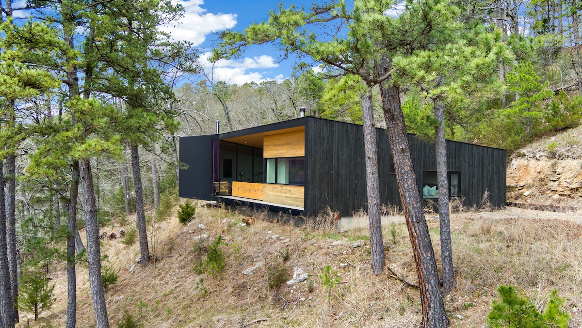 A modern cabin exterior is nestled among towering pine trees, showcasing a combination of dark siding and wooden accents. The structure features large windows that reflect the natural surroundings, providing an inviting connection to the landscape. A sloped area enhances the secluded feel.