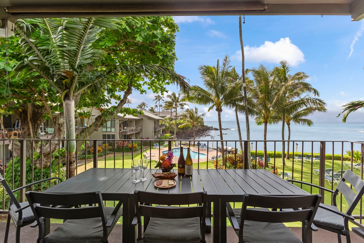 A dining table is set for four, featuring a fruit arrangement and wine, with an ocean view beyond. Lush palm trees and tropical landscaping frame the scene, while the serene waters of Honokeana Cove are visible in the distance.