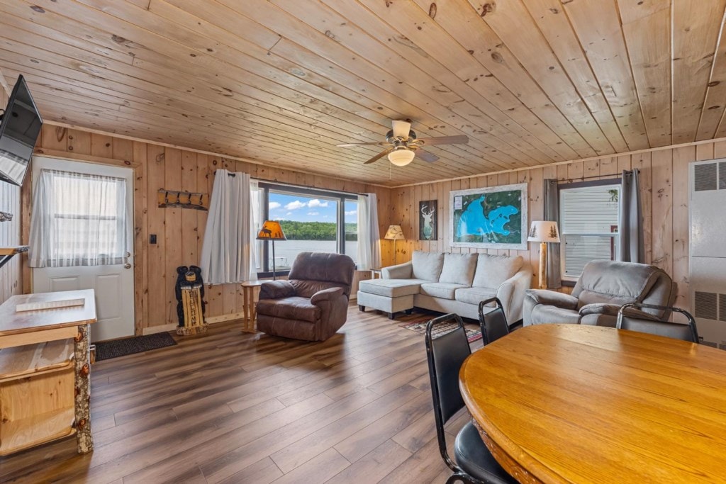 A spacious living area is featured, showcasing wood paneled walls and a warm wooden floor. A comfortable white sofa and armchair face a wall-mounted flat-screen TV. Large windows provide views of the outdoors, while a dining table with black chairs occupies the corner.