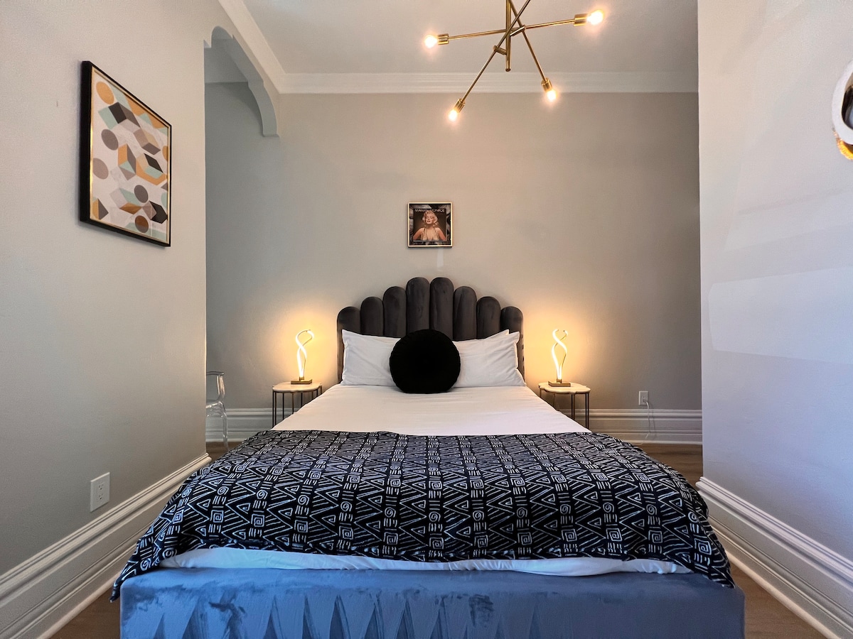 A cozy sleeping area features a plush headboard with a circular cushion in front of a queen-sized bed. Nightstands with unique lamps are positioned on either side, and a piece of wall art adds a touch of color to the neutral walls.