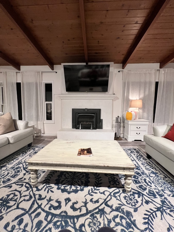 A cozy living area features a white marble fireplace with a flat-screen television mounted above it. Light-colored sofas are arranged around a coffee table, all set on a decorative blue area rug. Large windows draped with sheer curtains allow for natural light to enter the space.