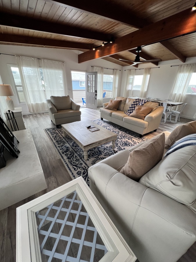 A bright living area features comfortable seating, including two armchairs and a spacious sofa. A coffee table is centrally placed on a patterned rug. Large windows allow natural light to fill the room, while sheer curtains provide a soft glow.