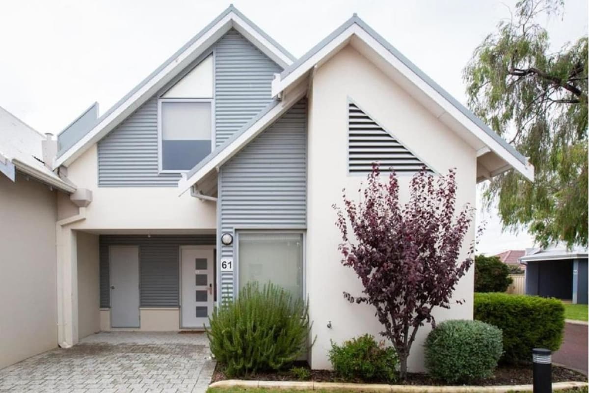 The exterior of the holiday home features a contemporary two-story design, with a combination of grey and white cladding. Lush shrubs and small trees border the entrance, while a paved driveway leads to the front door, creating an inviting approach to the property.