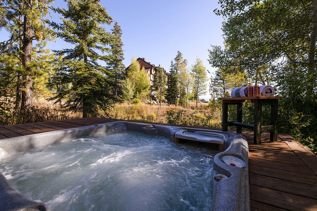 A private hot tub is displayed on a spacious deck, surrounded by trees, providing a serene outdoor space. A small table nearby holds rolled towels, adding to the relaxation ambiance. In the background, a multi-story building is partially visible among the foliage.