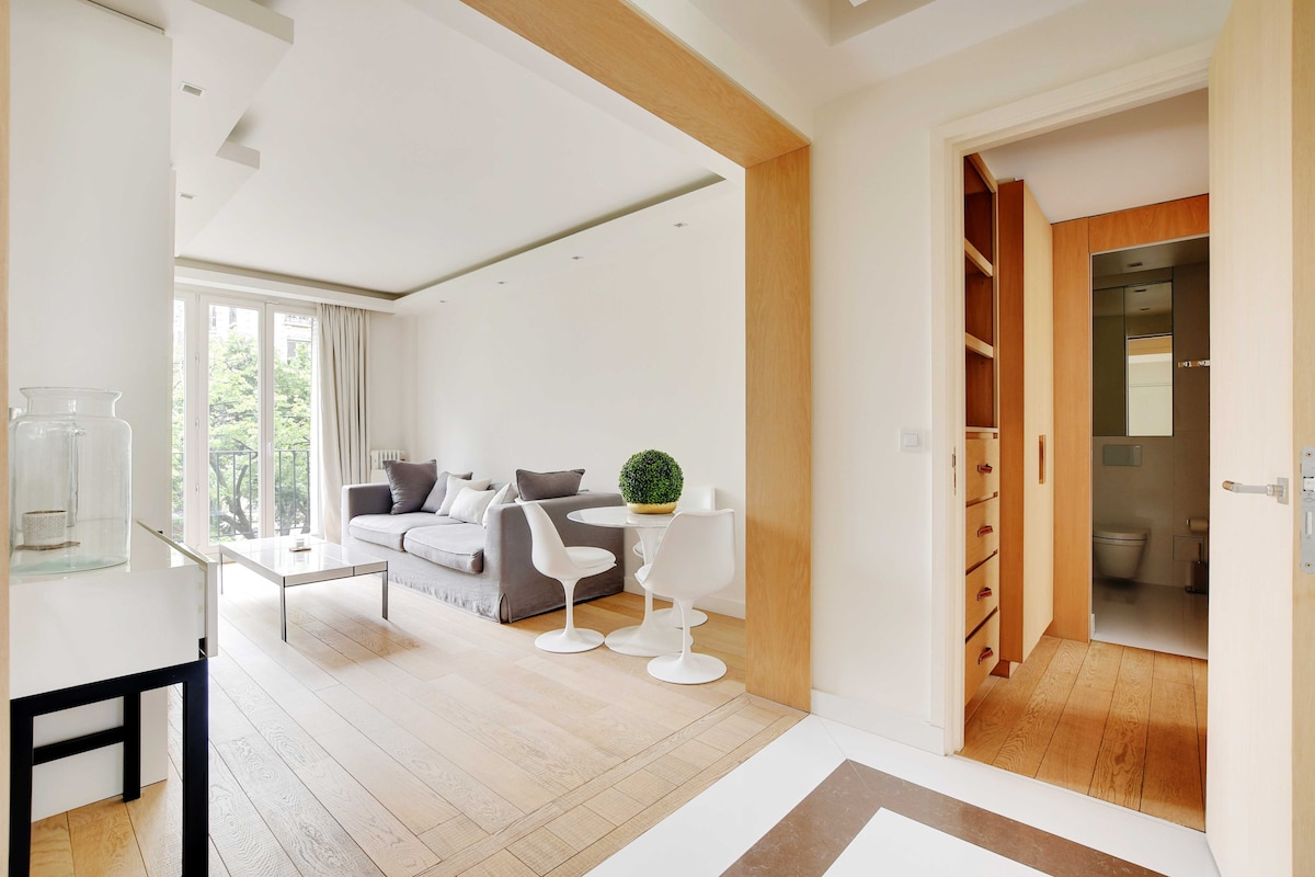 A modern living area features a large, comfortable sofa and a glass coffee table, enhanced by natural light from tall windows adorned with light curtains. A small decorative plant is placed on the table. An entryway leads to a bathroom with sleek fixtures and wooden storage options.