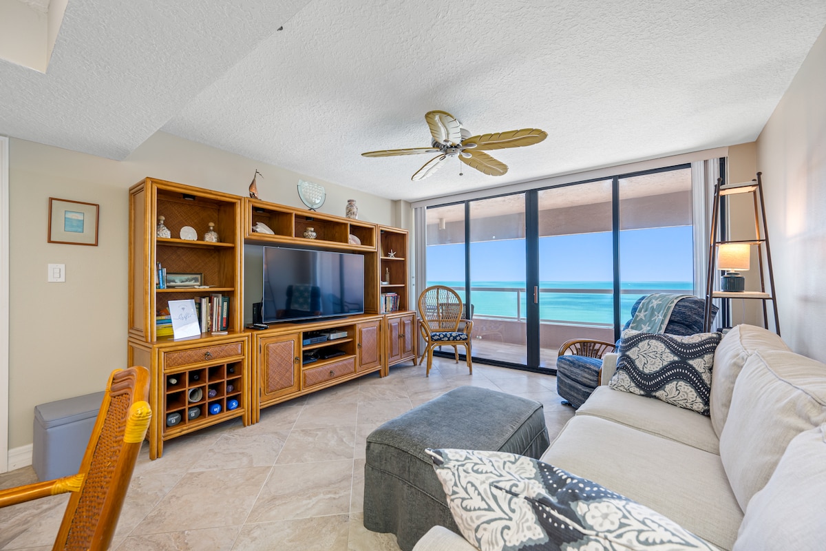 The living area features a spacious couch with coastal-themed cushions, a wooden entertainment unit with a flat-screen television, and a decorative bookshelf. A ceiling fan contributes to the airy ambiance, while large sliding glass doors offer unobstructed views of the ocean.