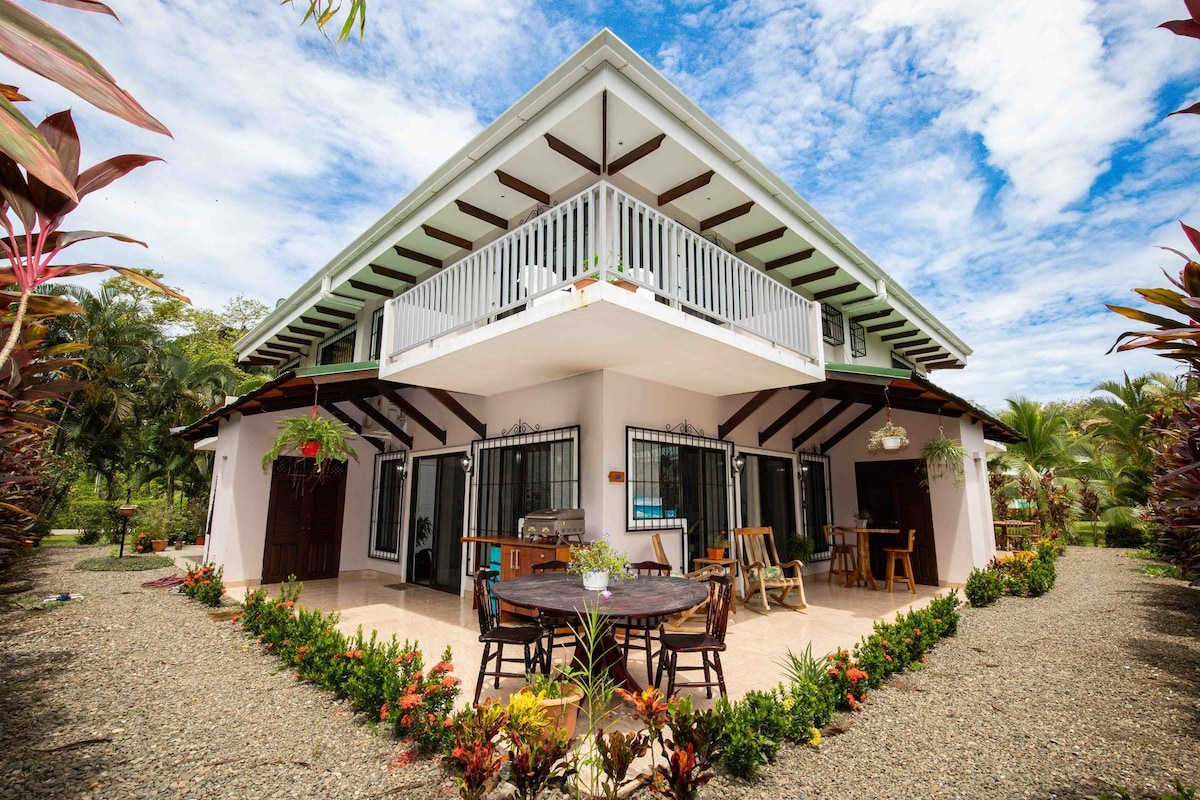 The two-story corner condo is surrounded by lush greenery, featuring a spacious patio area with outdoor seating. The structure includes a balcony on the second floor, and large windows offer views of the well-maintained garden. A tranquil atmosphere is created by the vibrant landscaping.
