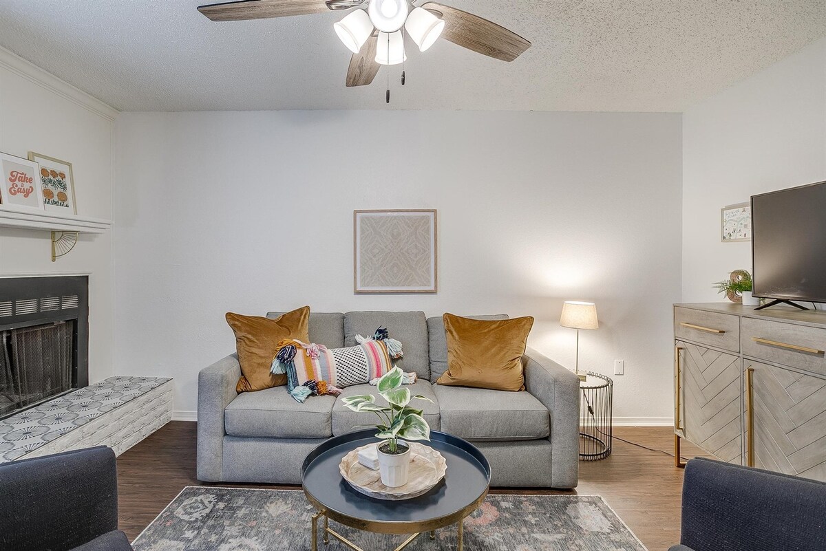 A cozy living room is presented with a spacious gray couch adorned with decorative pillows and a throw. A circular coffee table holds a potted plant, while a small lamp provides soft lighting. A television is positioned on a side cabinet, and a decorative fireplace adds character.