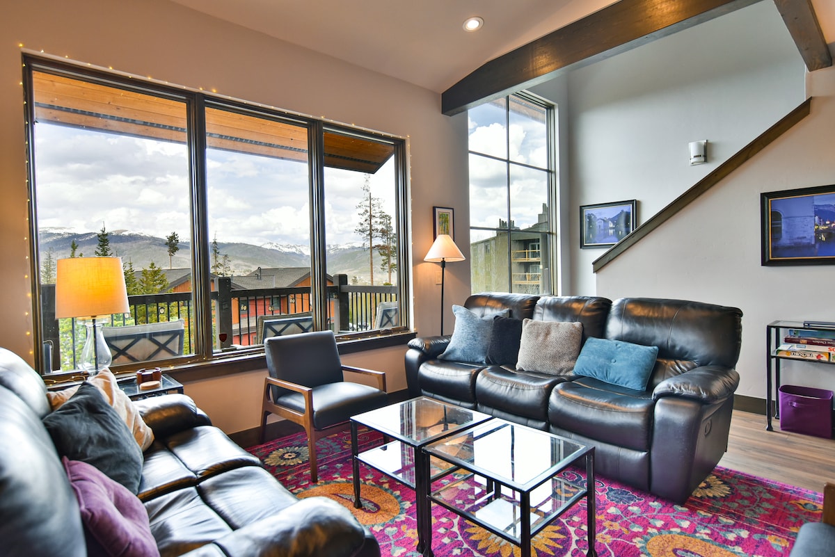 A cozy living area features a comfortable leather sofa and armchair, framed by large windows offering sweeping views of the mountains. A glass coffee table sits on a colorful area rug, complemented by ambient lighting from a standing lamp.