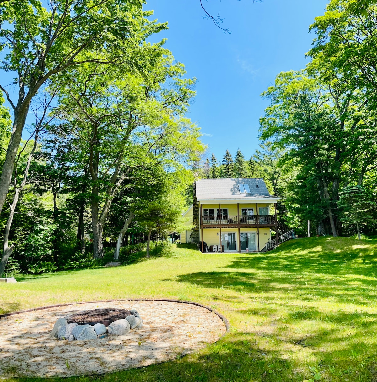 A sunny outdoor view showcases the cottage amid lush trees, framed by a spacious lawn. A fire pit surrounded by smooth stones is set in the foreground, inviting gatherings. The two-story structure features large deck and glass doors on the lower level, enhancing the natural setting.