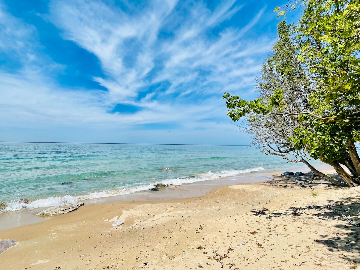 A serene beach scene is depicted, featuring soft sand meeting clear, turquoise waters. Gentle waves roll onto the shore, where sparse rocks are visible. A few trees provide shade along the coastline, and a lightly clouded sky creates a tranquil ambiance.
