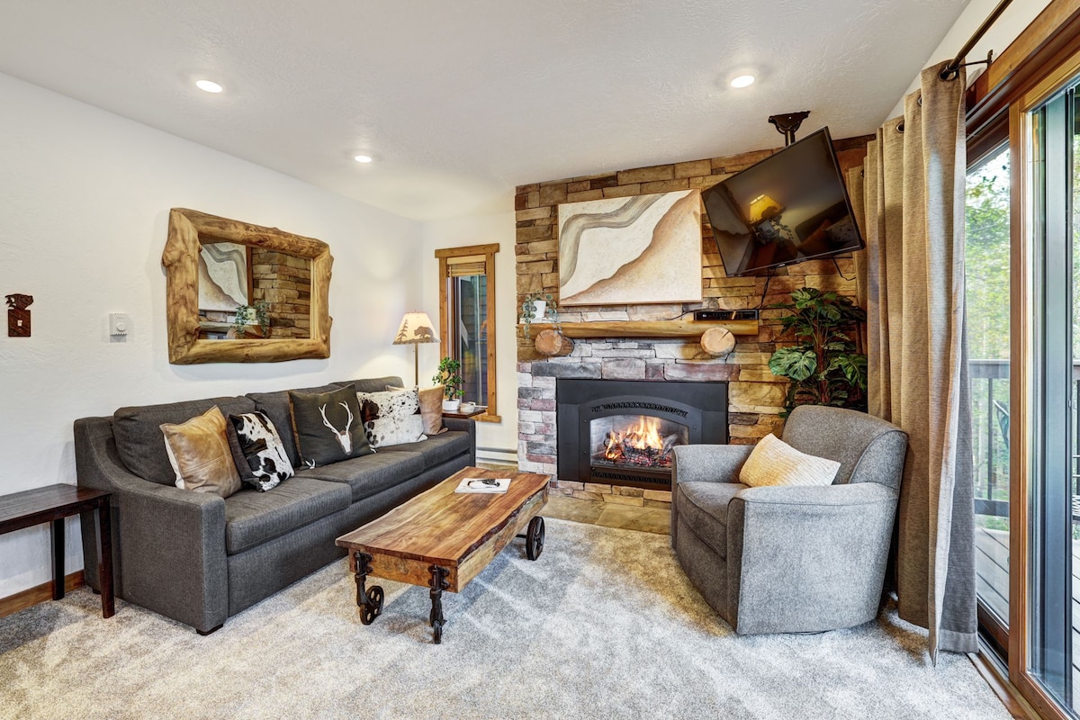 A cozy living room is furnished with a charcoal gray sectional sofa and a single armchair, accented by a rustic wooden coffee table. A flat-screen TV is mounted above a stone fireplace, surrounded by artwork, while natural light enters through large sliding glass doors leading to the back deck.