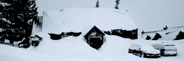 A snow-covered exterior of a mountain lodge is visible, with the sloped roof laden with thick snow. The entrance is partially obscured by the snow drifts, and vehicles are parked outside, surrounded by a winter landscape.