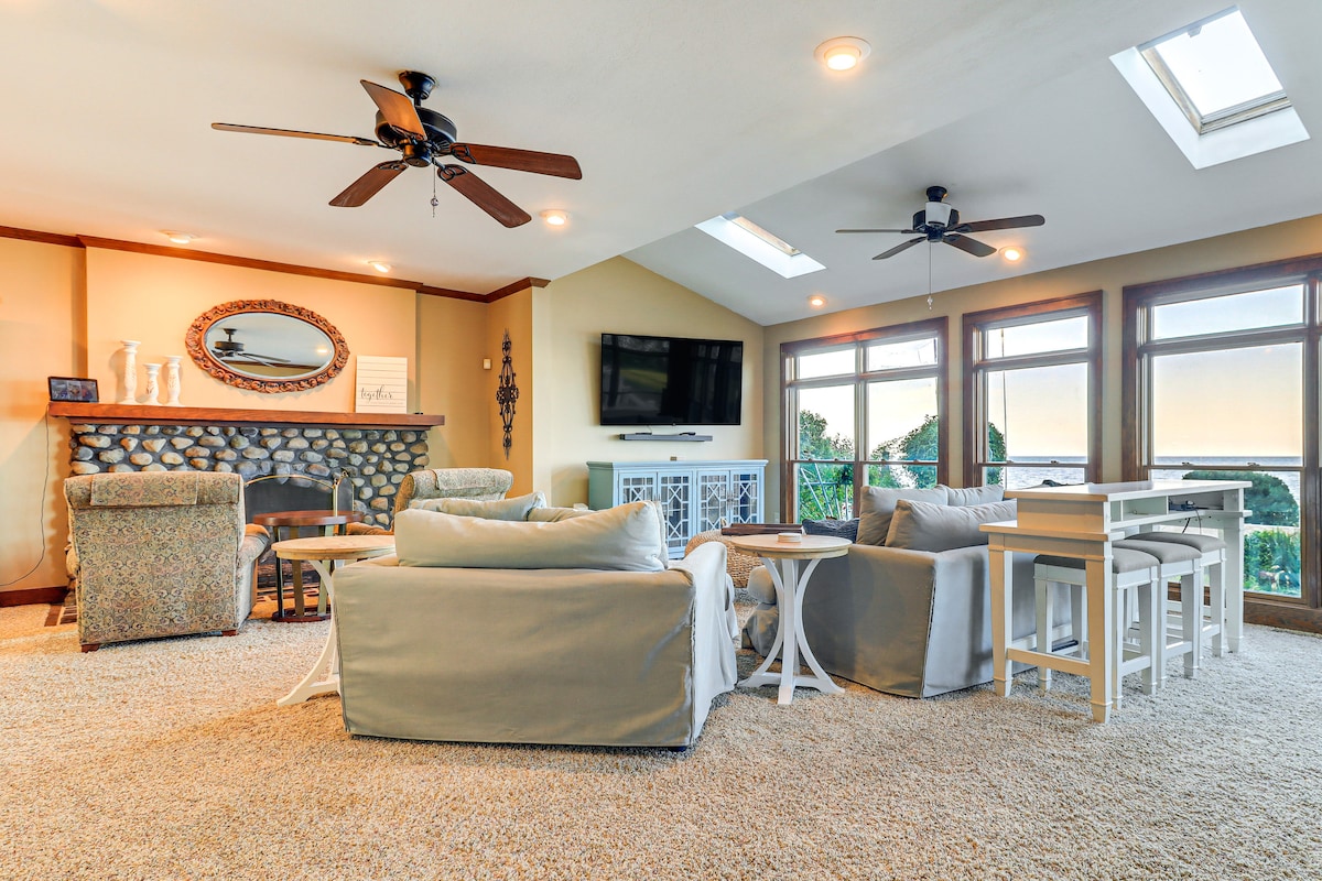 A spacious living area is highlighted by large windows allowing natural light and scenic views. Comfortable seating, including plush sofas and a cozy armchair, surrounds a coffee table. A flat-screen TV is mounted above a stone fireplace, complemented by a dining table nearby.