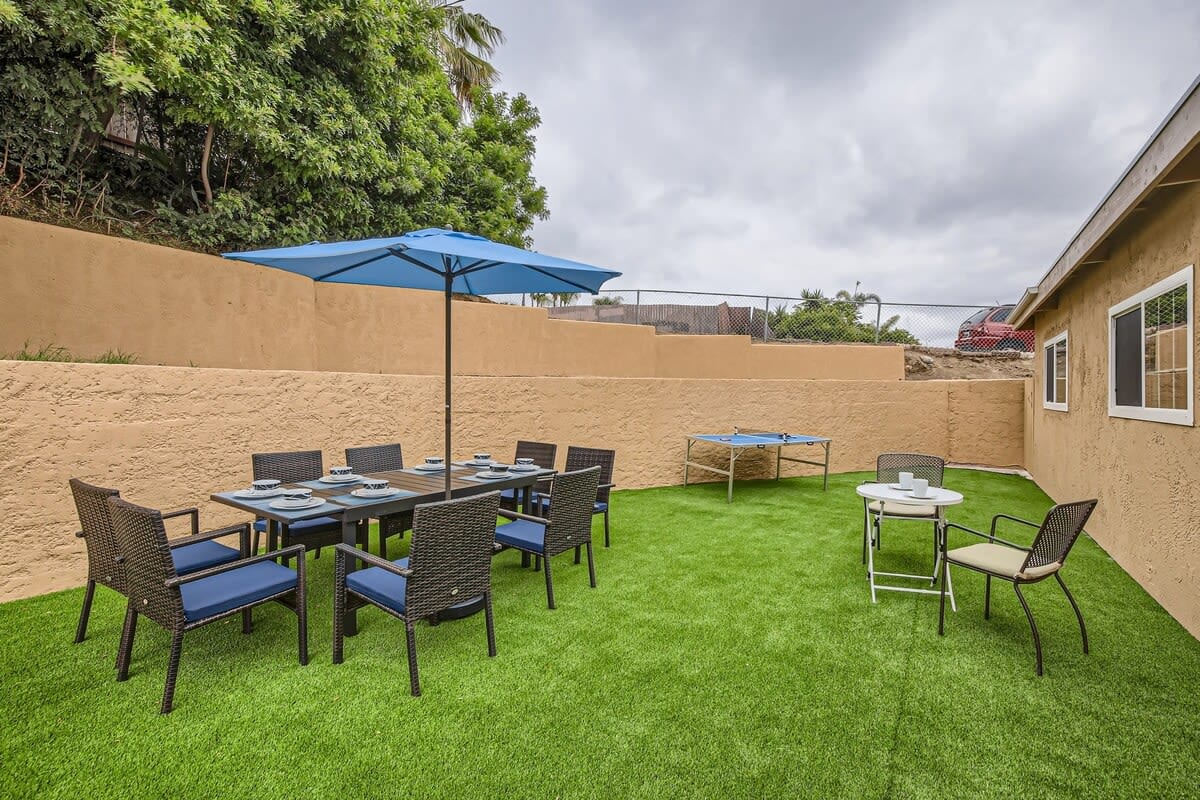 An outdoor space features a well-maintained artificial lawn surrounded by stucco walls. A dining table shaded by a blue umbrella is surrounded by black chairs, while a Ping Pong table is positioned nearby for recreational activities.