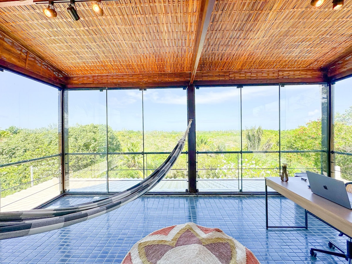 A spacious room is designed with large glass walls that offer expansive views of the surrounding greenery. Inside, a hammock is suspended for relaxation, complemented by a modern workspace with a desk and computer. Natural light floods the area, enhancing the connection with nature.