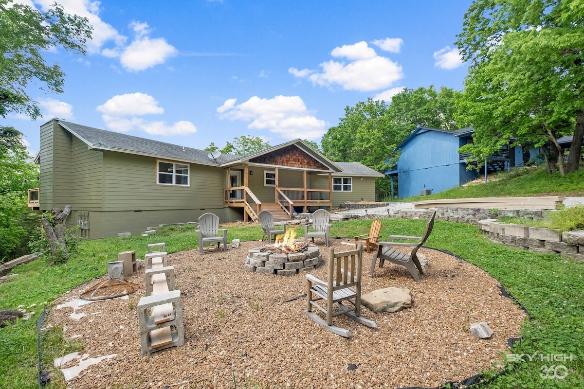 A firepit area is surrounded by wooden seating arranged on a circular stone base. The backyard includes a mix of gravel and grass, with trees providing greenery against a clear blue sky. The inviting exterior of the home is visible in the background.