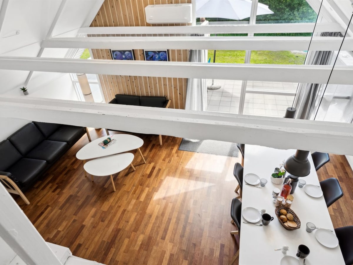 The open-plan living and dining area features wooden flooring and large windows allowing natural light to fill the space. A comfortable black sofa and a white coffee table are positioned near the dining table, set with dishes and a beverage station.