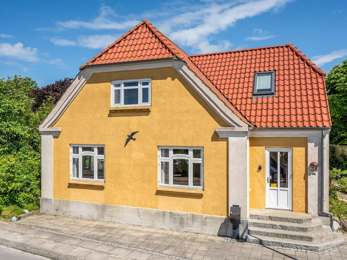 The exterior of a yellow house features a red tiled roof and multiple windows. The entrance is framed by a few steps leading up to a welcoming front door. Lush greenery surrounds the property, complementing its inviting appearance.