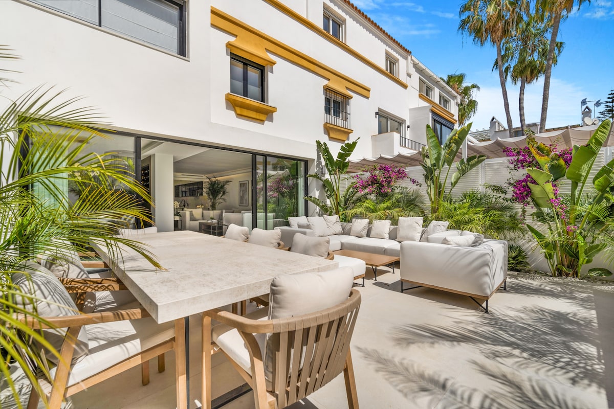 An outdoor terrace showcases a large dining table surrounded by comfortable armchairs. Lush greenery and tropical plants enhance the space, providing a serene atmosphere. Sliding glass doors lead into the living area, inviting natural light throughout the area.