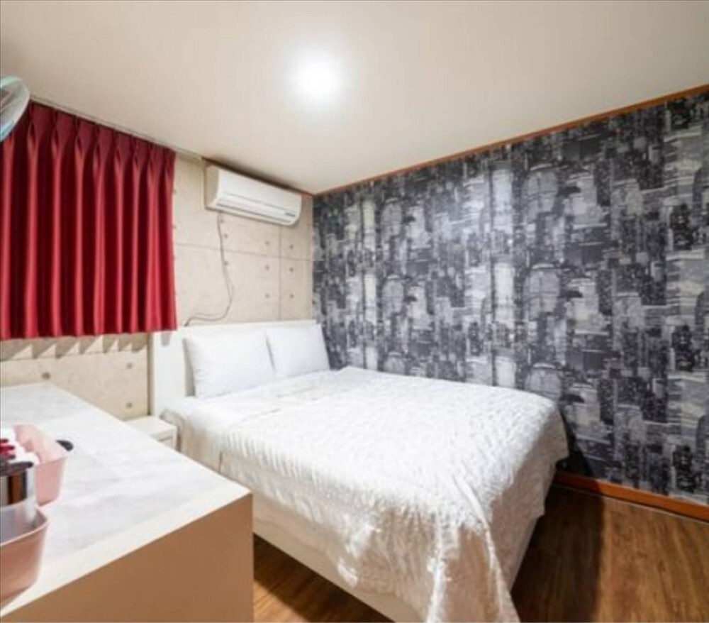 A neatly arranged sleeping area features a spacious bed dressed in white linens. A patterned accent wall adds visual interest, while red curtains frame a window. An air conditioning unit is mounted above the bed, providing comfort during warmer days.