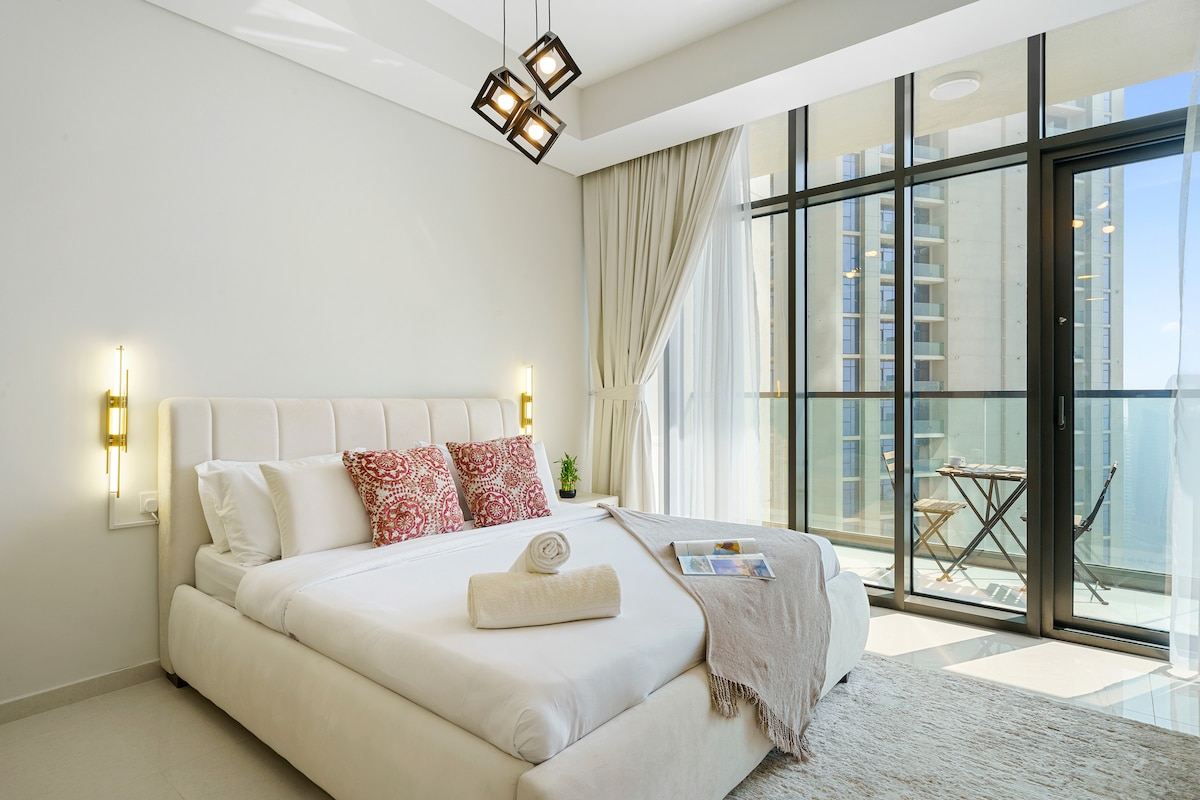 A modern studio apartment showcases a spacious bed with plush pillows, complemented by soft lighting fixtures. Large glass doors lead to a private balcony, offering views of the surroundings. Light-colored curtains and a cozy throw enhance the inviting atmosphere of the room.
